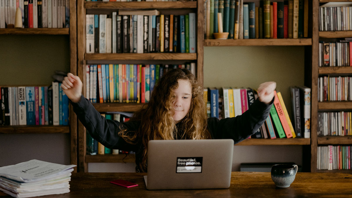Person at a laptop in front of bookshelves raising both fists in triumph