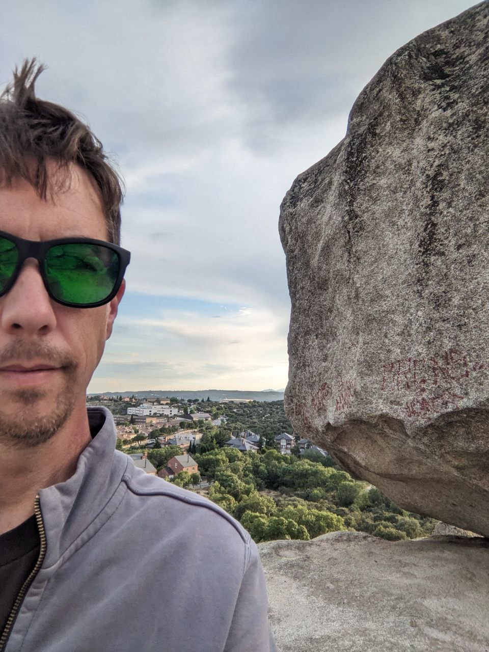 Man in green sunglasses taking a selfie beside a large granite boulder with a village below