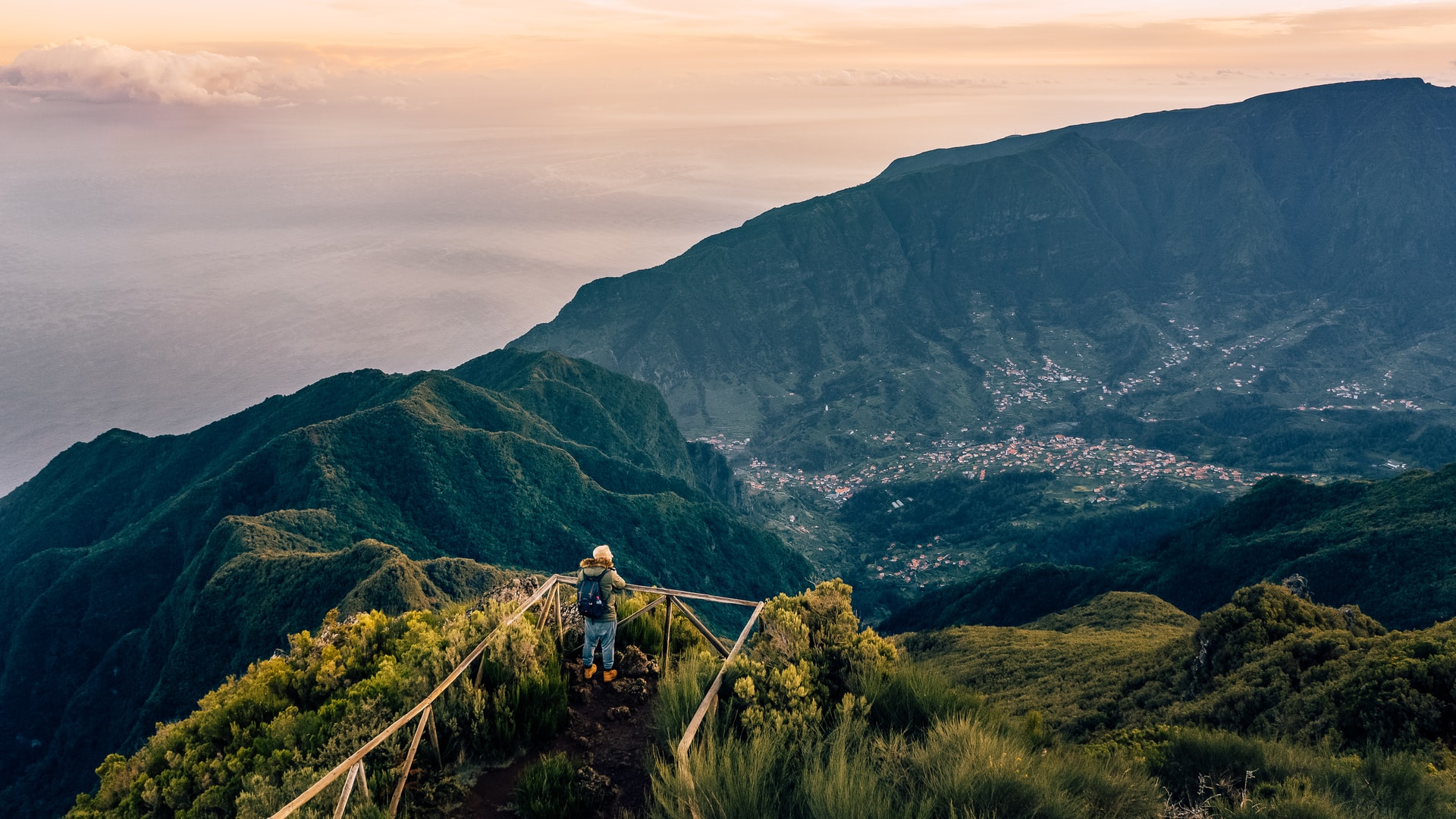 Hiker standing at a railing viewpoint above green mountains and a valley village