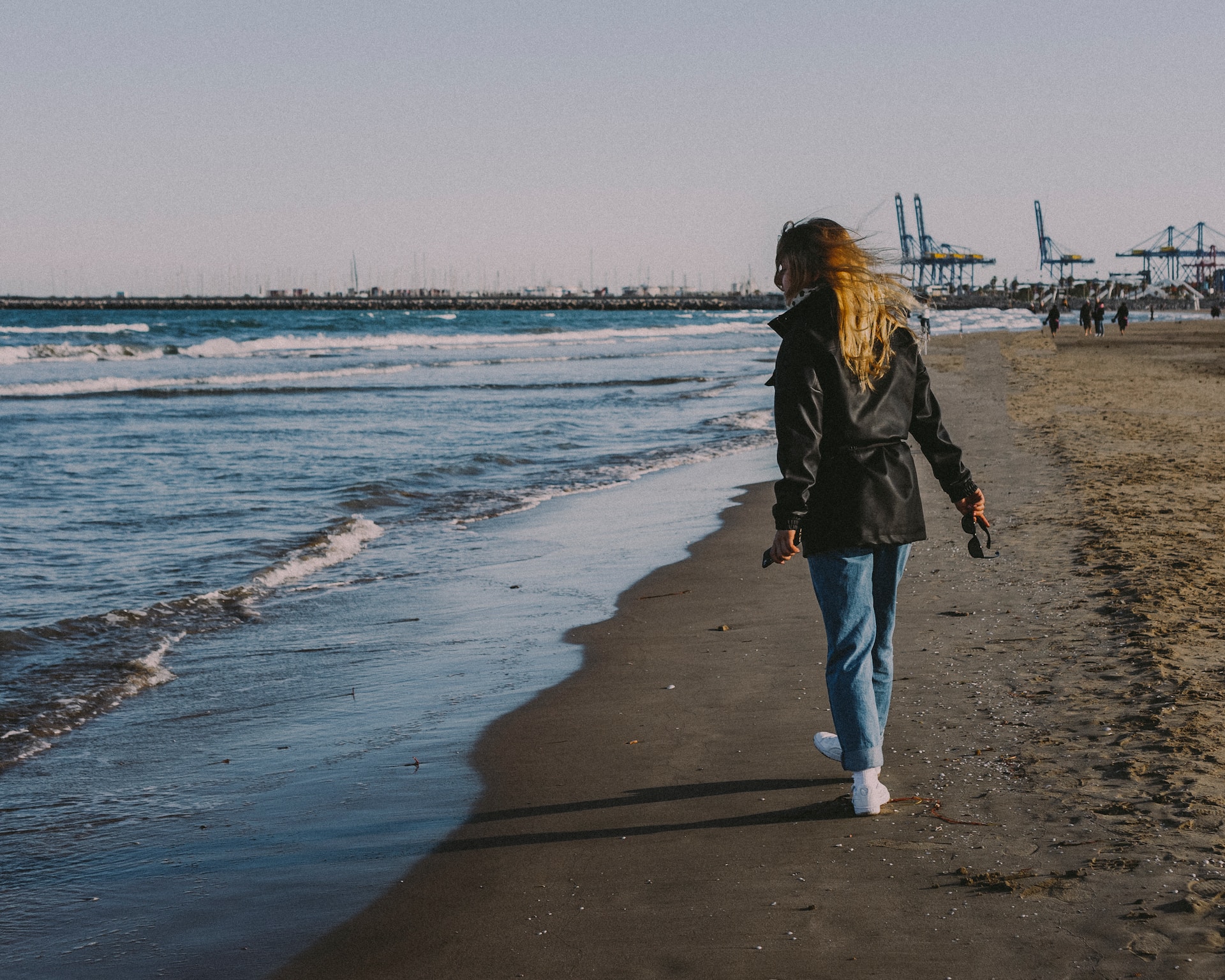 Woman walking along a sandy beach at the water's edge, port cranes in the distance